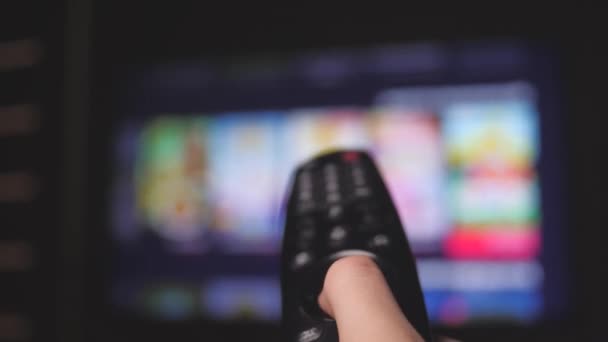 Woman hand selects internet tv channels with remote control, close-up. Person controls TV using a modern remote control. Girl watches smart TV and uses black remote control. Blurry tv scrolls pages - Footage, Video
