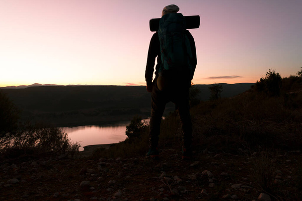 Silueta de un hombre senderista en vista trasera al anochecer cerca de un lago y en el fondo unas montaas con el sol ocultandose. Esztilo de vida.  - Fotó, kép
