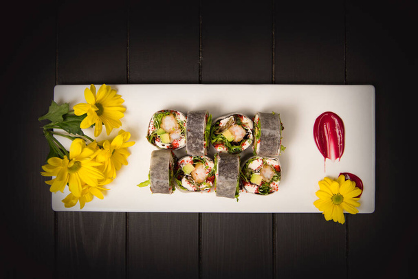 Draufsicht auf gesunde japanische Sushi-Rolle ohne Reis mit Tempura-Garnelen in Daikon-Rettich gewickelt. Gelbe Kamillenblüten malen Pinselstrich-Dekorationen auf weißem rechteckigen Teller. Schwarzer Hintergrund - Foto, Bild