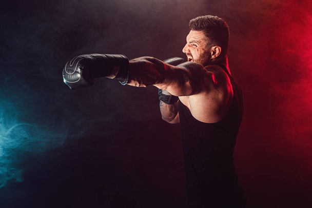 Bearded tattooed sportsman muay thai boxer in black undershirt and boxing gloves fighting on dark background with smoke. - Фото, изображение