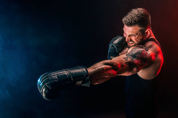 Bearded tattooed sportsman muay thai boxer in black undershirt and boxing gloves fighting on dark background with smoke. - Fotoğraf, Görsel