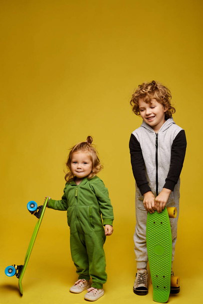 Happy curly kid and cute little girl in kigurumi holding longboards and posing on the yellow background, isolated. Two smiling kids in summer outfits with skateboards on yellow. Kids fashion - Photo, Image