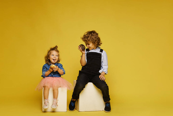 Cute children eat delicious donuts over a yellow background. Cute little girl and curly boy in summer outfits eat tasty pastry on the yellow background, isolated with copy space - Photo, image