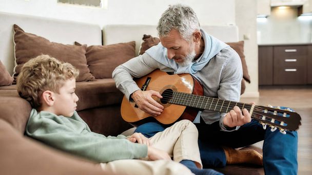 Transmitting knowledge. Grandfather teaching grandson how to play guitar while spending time together at home sitting in the living room - Foto, Imagem