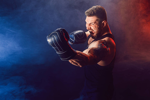 Bearded tattooed sportsman muay thai boxer in black undershirt and boxing gloves fighting on dark background with smoke. - Photo, Image