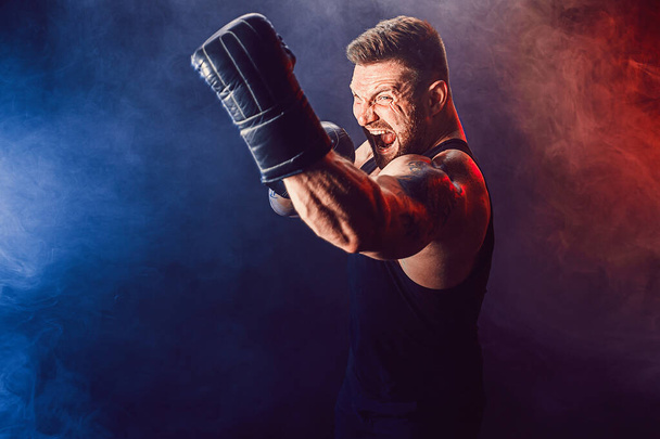 Bearded tattooed sportsman muay thai boxer in black undershirt and boxing gloves fighting on dark background with smoke. - Фото, изображение