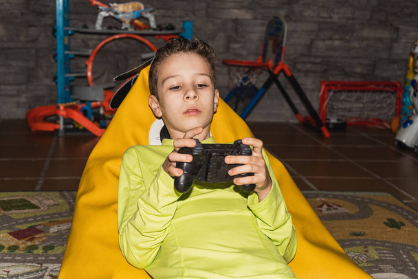 Child playing video games lying on a pouf wearing a yellow T-shirt with the controller in his hand. Lifestyle - Fotoğraf, Görsel