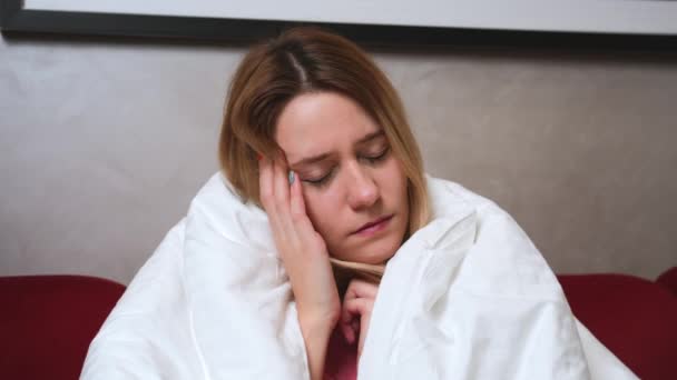 jong blank ziek meisje met covid of griep zitten onder deken op de bank in de woonkamer en hebben hoofdpijn. Zieke vrouw met coronavirus symptomen. - Video