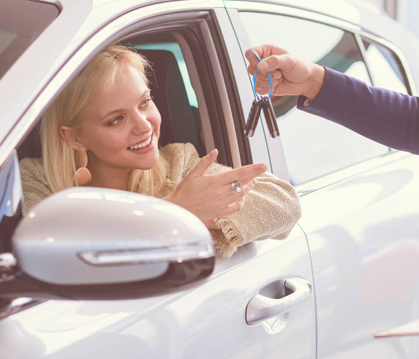 Beautiful young woman is getting key and smiling while sitting in a new car in dealership - 写真・画像