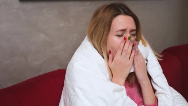 fiatal kaukázusi beteg lány, megfázással vagy megfázással, aki a kanapé alatt ül a nappaliban és szalvétában köhög. Beteg nő koronavírus tünetekkel. - Felvétel, videó