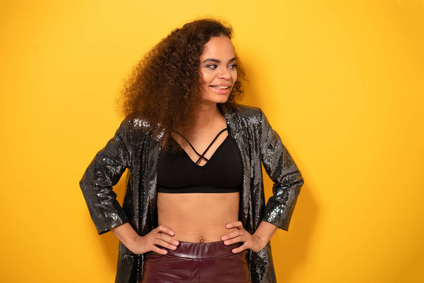 Happy Afro American girl with beautiful hairstyle posing smile looking side away with hand on hips wearing shiny black jacket and black top. Happy young girl fixing hair standing on yellow background - Foto, afbeelding