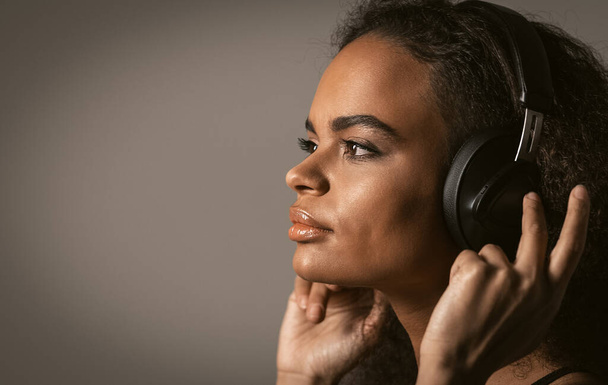 Charming girl portrait of an African-American looking sideways listening music wearing headphones holding it with hands, isolated on grey background, emotionally move, have fun. Close up - Photo, image