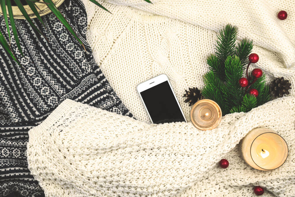 Современный зимний уютный фон, шерстяной и вязаный свитер, еловая ветвь с красными ягодами и свечами, холостой смартфон - Фото, изображение