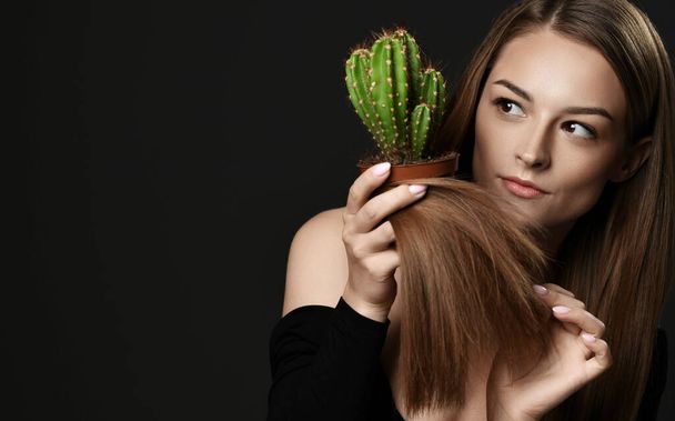 Young woman with long silky straight hair compares hair split ends with cactus plant in pot she holds looks aside - Photo, Image