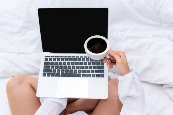 Close up hand holding cup coffee drink on white bed in the bedroom. Women working on laptop and drinking coffee in morning relax mood in winter season. Lifestyle Concept. - Foto, Imagem