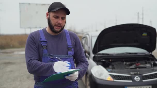 mannelijke automonteur bij een kapotte auto op de weg. Vertelt over storingen en maakt aantekeningen in een notebook. - Video