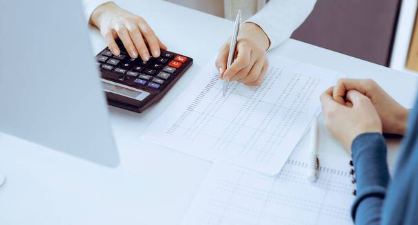 Accountant checking financial statement or counting by calculator income for tax form, hands close-up. Business woman sitting and working with colleague at the desk in office. Audit concept - Фото, изображение
