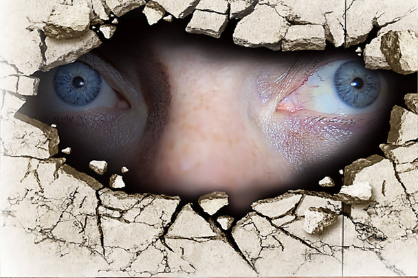 De blauwe ogen van een man die door een scheur in de muur kijkt. - Foto, afbeelding