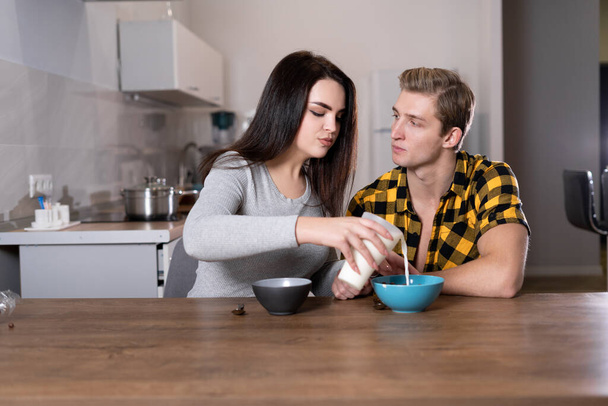 joven pareja caucásica desayunando en casa. Una mujer vierte leche para su marido en un tazón de cereal. - Foto, Imagen