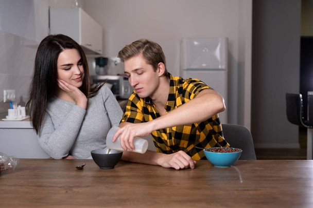 young Caucasian couple having breakfast at home. a man pours milk of his beloved into a bowl of cereal. - Photo, Image