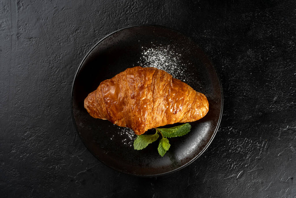 Golden crispy croissant on a black plate, garnished with mint leaves and powdered sugar. Fresh bakery. Dessert for breakfast. - Fotó, kép
