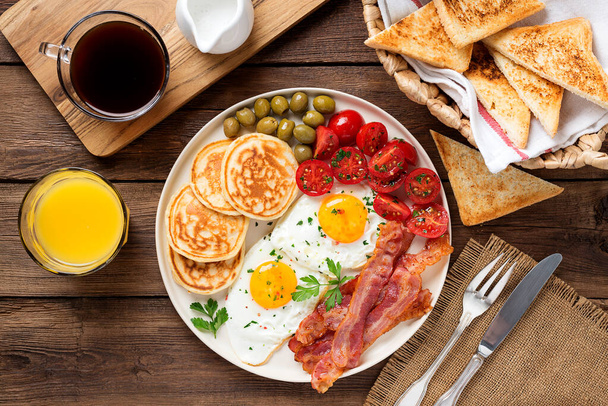 Американский завтрак с двумя жареными яйцами, блинами, салатом из помидоров, беконом и зелеными оливками. вид сверху - Фото, изображение