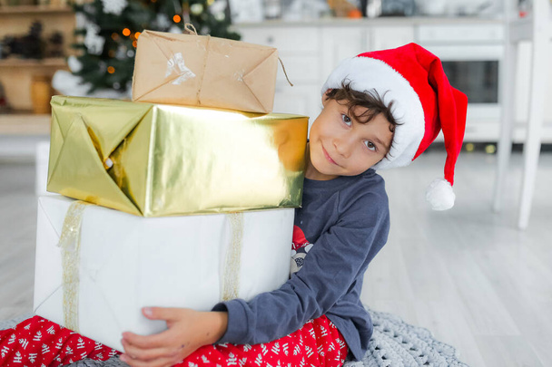 curly haired boy in santa claus hat holding gifts at home with christmas decor - Foto, afbeelding