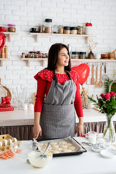 Walentynki. Kobieta w czerwonej sukience i szarym fartuchu robi walentynkowe ciasteczka w kuchni - Zdjęcie, obraz