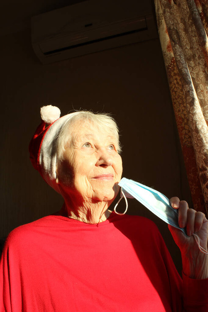 Senior woman near the window in the sunny morning with a Santa hat wears a medical mask due to the coronavirus. Self isolation away from everyone to avoid contagion - Photo, Image