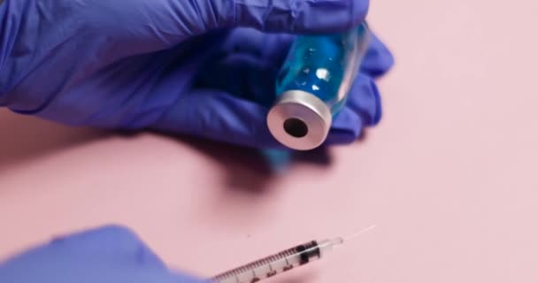 Vrouwen hand in hand blauwe latex handschoen houdt spuit injectie en glazen fles met vloeibaar vaccin op roze achtergrond - Video
