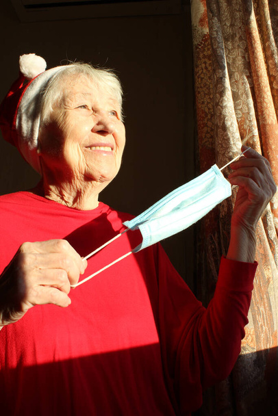 Старша жінка біля вікна в сонячний ранок з капелюхом Санта носить медичну маску через коронавірус. Самоізоляція від усіх, щоб уникнути зараження
 - Фото, зображення