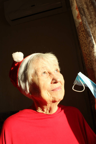 Starsza kobieta przy oknie w słoneczny poranek w czapce Mikołaja nosi maskę medyczną z powodu koronawirusu. Samoizolacja z dala od wszystkich, aby uniknąć zakażeń - Zdjęcie, obraz