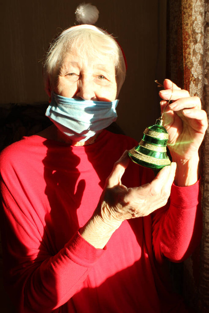 Senior woman near the window in the sunny morning with a Santa hat wears a medical mask due to the coronavirus. Self isolation away from everyone to avoid contagion - Photo, Image