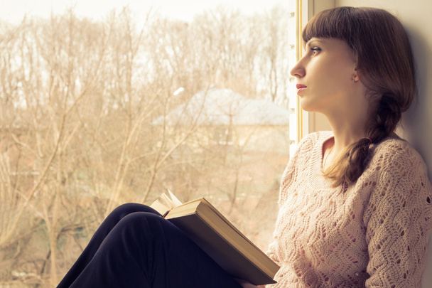 Młoda dorosła dziewczyna czyta książkę przy oknie - Zdjęcie, obraz