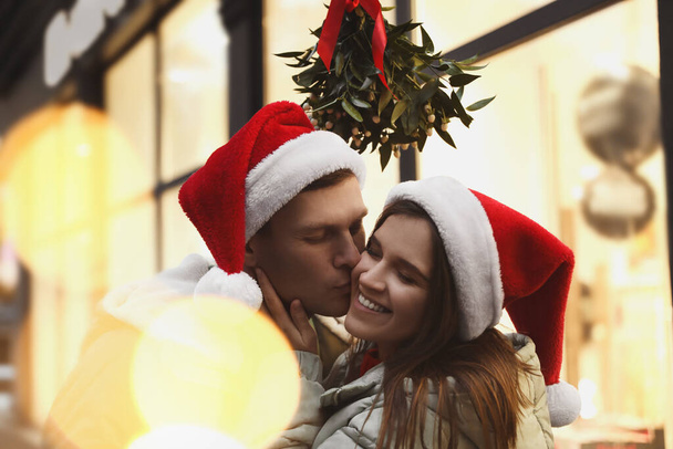 Happy man kissing his girlfriend under mistletoe bunch outdoors, bokeh effect - Fotó, kép