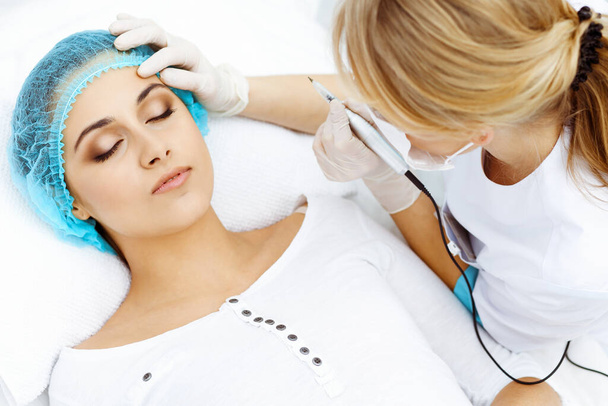 Professional beautician doing eyebrow tattoo at woman face. Permanent brow makeup in beauty salon, closeup. Cosmetology treatment - Фото, зображення