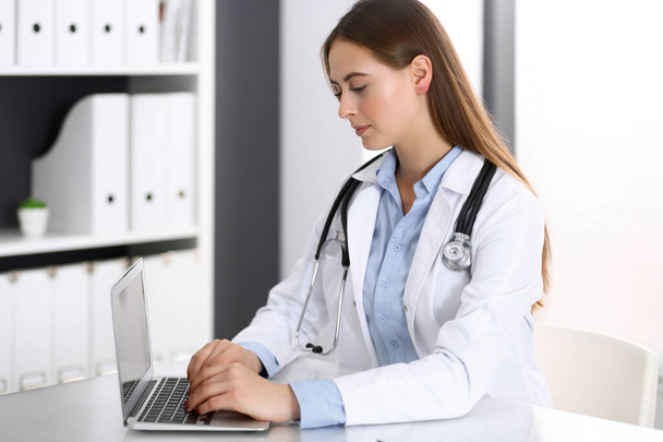 Doktor pisze na laptopie, siedząc przy biurku w szpitalu. Lekarz w pracy. Koncepcja medycyny i opieki zdrowotnej - Zdjęcie, obraz