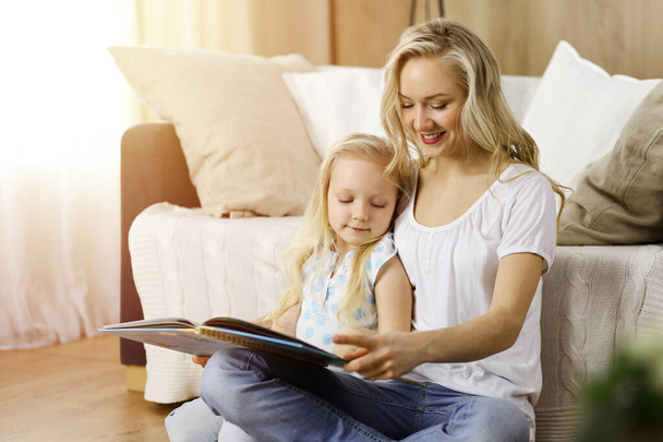 Happy family. Blonde young mother reading a book to her cute daughter while sitting at wooden floor in sunny room. Motherhood concept - Foto, Bild