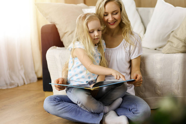 Happy family. Blonde young mother reading a book to her cute daughter while sitting at wooden floor in sunny room. Motherhood concept - Fotó, kép