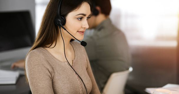 Call center operators at work. Two young people in headsets are talking to the clients, while sitting in sunny office - Valokuva, kuva