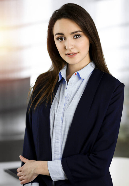 Intelligent young business woman in a blue jacket is standing at her workplace. Portrait of a specialist in sunny office - Foto, imagen
