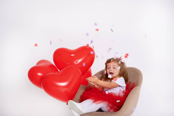 maluch dziewczyna na czerwony spódnica siedzi w krzesło rzuca confetti na biały tło - Zdjęcie, obraz