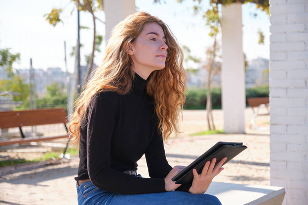 Kızıl saçlı kadın, parkta kitap okuyor ya da tablet veya e-kitapta çalışıyor. Dijital göçebe girişimci konsepti. - Fotoğraf, Görsel