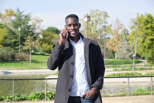 Joven hombre negro africano hablando y sonriendo en el teléfono inteligente mirando a la cámara en un parque. Empresario al teléfono. - Foto, Imagen