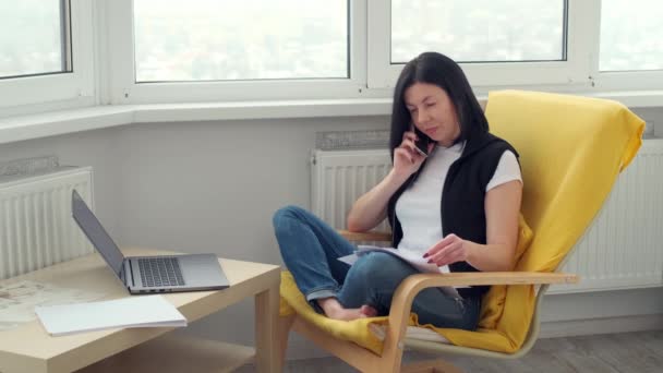 nainen rento vaatteet istuu nojatuoli huoneessa, työskentely kannettavan puhelimen ja asiakirjoja. etäisyyttä verkossa työtä kotona. Brunette liikenainen tekee freelance Internetissä - Materiaali, video