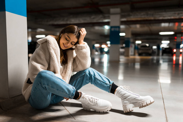 Fashionable girl listening to music in wireless headphones while sitting in the underground parking of shopping mall - Foto, imagen