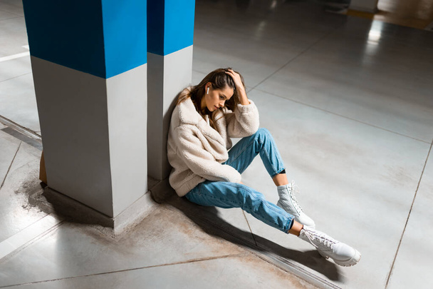Модная девушка слушает музыку в беспроводных наушниках, сидя на подземной парковке торгового центра - Фото, изображение