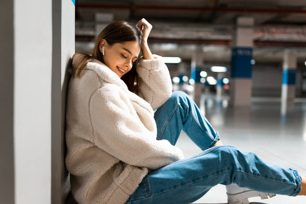 Fashionable girl listening to music in wireless headphones while sitting in the underground parking of shopping mall - 写真・画像