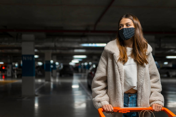 Elegante ragazza in maschera medica con carrello della spesa nel parcheggio sotterraneo del centro commerciale - Foto, immagini