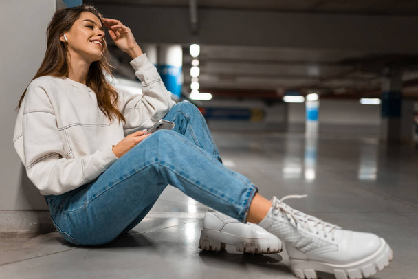 Stylish girl in the underground parking sits on the floor and listens to music on wireless headphones from smartphone - Фото, зображення